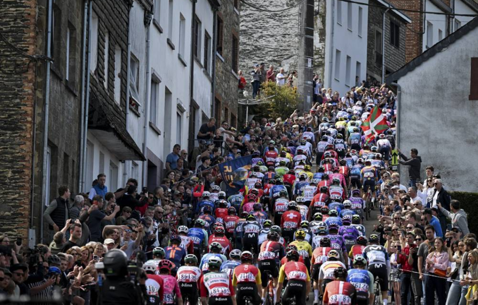 Grande torcida de ciclismo na rua estreita com muitos ciclistas em disputa e público vibrando, na colina de uma cidade antiga com moradores e turistas assistindo.