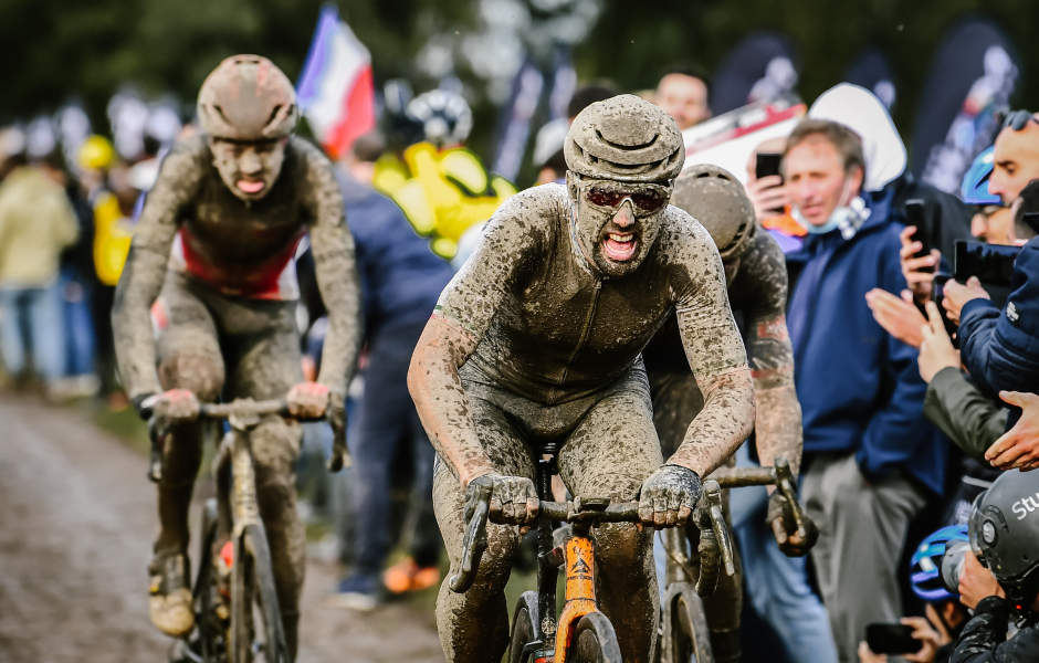 Participantes de uma competição de ciclismo estilo maratona, cobertos de lama, demonstrando esforço e determinação, com público assistindo e fotografando ao redor.