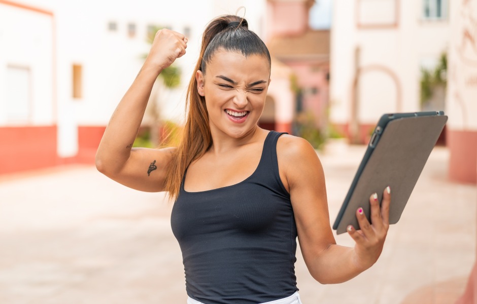 Mulher jovem comemora vibrante ao olhar para um tablet em ambiente externo. A imagem expressa sucesso, conquista de metas e felicidade com resultados digitais.