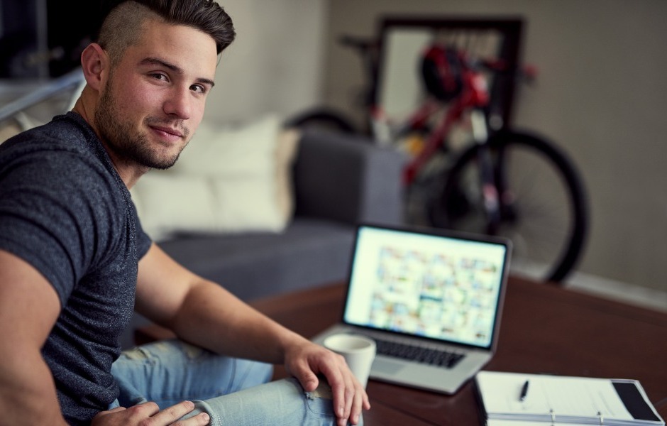 Homem sentado em um sofá olhando para a câmera, com um notebook aberto e uma bicicleta ao fundo.