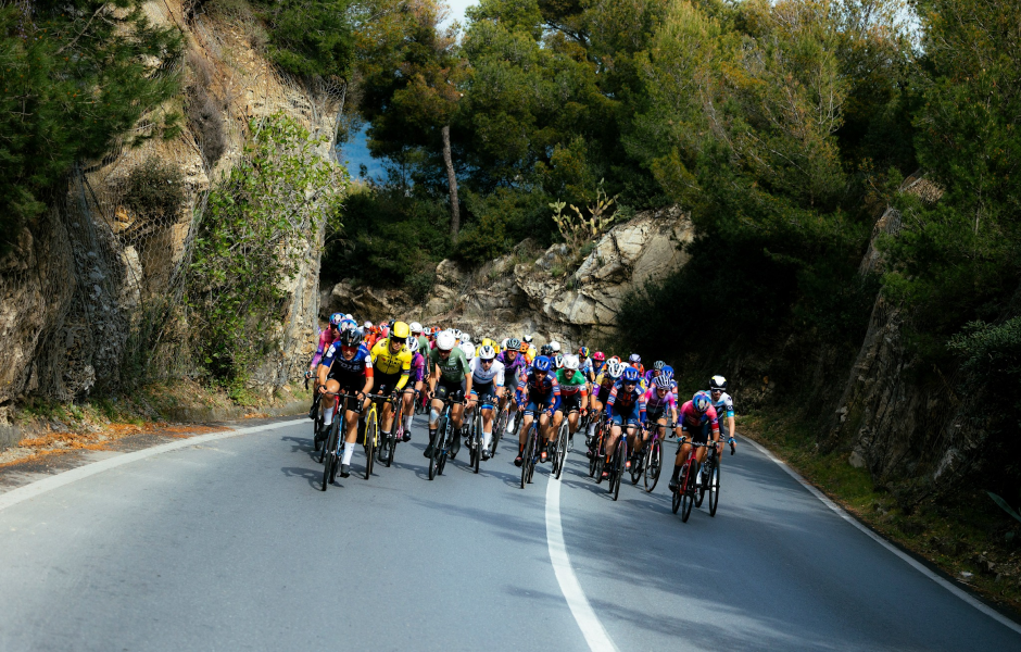 Grupo de ciclistas pedalando em uma estrada cercada por natureza, com árvores e rochas ao redor, durante um passeio ao ar livre, promovendo o ciclismo.