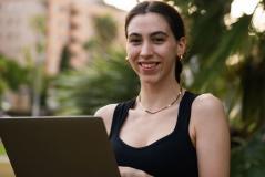 Jovem mulher sorridente usando blusa preta e segurando um tablet em ambiente ao ar livre, com vegetação e edifícios ao fundo.