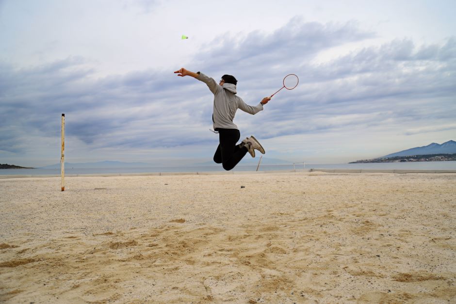 Jogador homem em posição de ataque no ar, dentro de quadra de areia à beira-mar, ele está prestes a acertar peteca de beach badminton