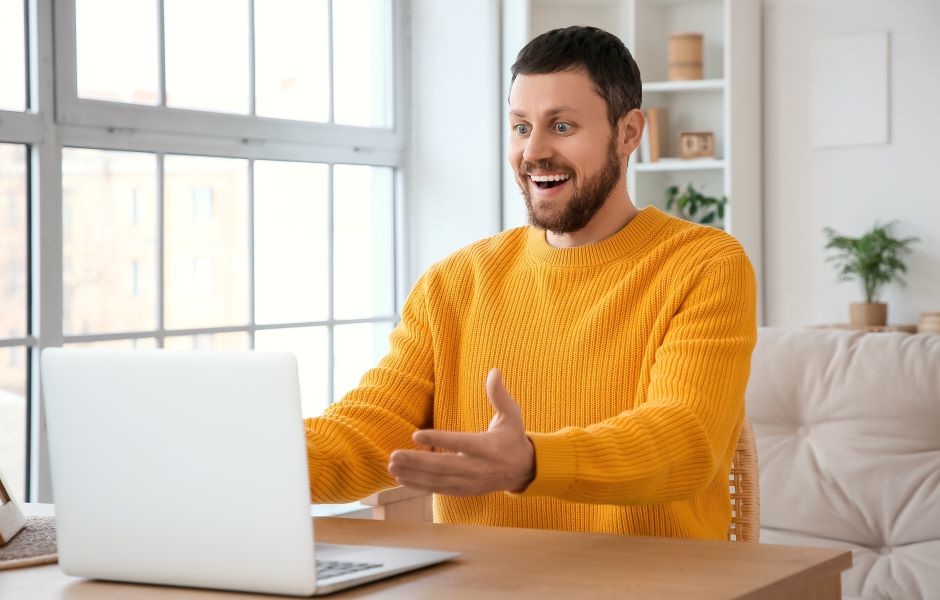 Homem alegre com barba e cabelo curto, usando suéter amarelo, interagindo por vídeo chamada em um ambiente bem iluminado com grande janela de fundo.