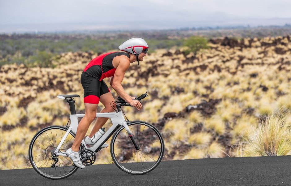 Homem praticando ciclismo em um ambiente natural desértico, vestindo roupa esportiva vermelha e capacete branco, pedalando em uma estrada pavimentada com vista de vegetação ao fundo.