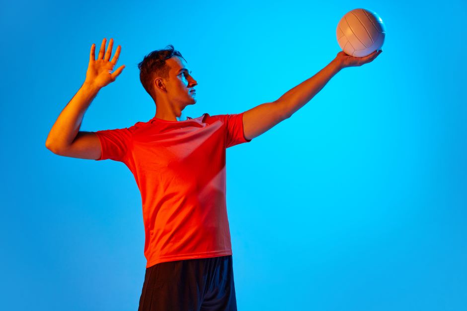 Homem posicionado para efetuar um saque de vôlei, um de seus braços estendidos com um bola de vôlei em suas mãos