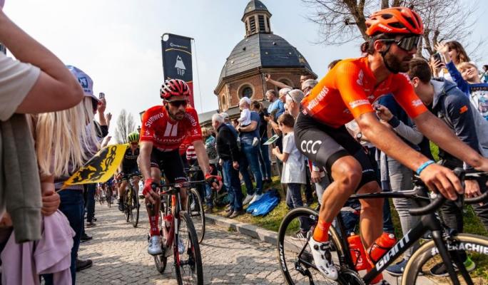 Atletas profissionais de ciclismo competindo em uma corrida oficial com uma multidão de espectadores apoiando ao lado de uma igreja histórica ao ar livre.