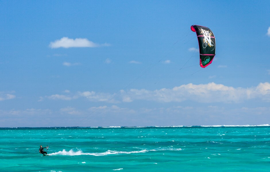 Imagem de um praticante de kitesurf deslizando sobre águas turquesas, utilizando uma pipa para ser impulsionado pelo vento.