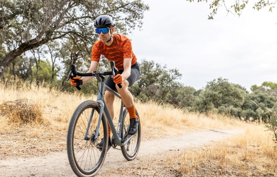 Pessoa pedalando de bicicleta na natureza com capacete, óculos de proteção, camiseta laranja, calções e meias laranja, aproveitando um passeio ao ar livre em meio a árvores e caminho de terra.