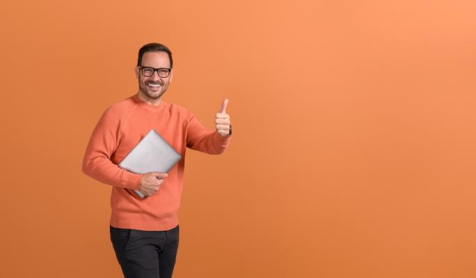 Homem sorridente vestindo um suéter laranja, segurando um laptop e mostrando sinal de positivo, em fundo laranja.