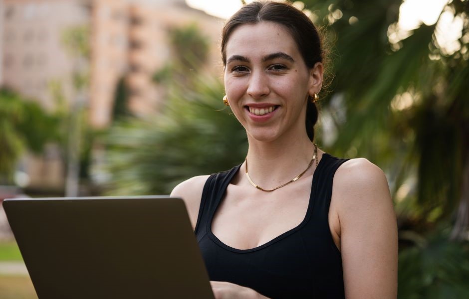 Jovem mulher sorridente usando blusa preta e segurando um tablet em ambiente ao ar livre, com vegetação e edifícios ao fundo.