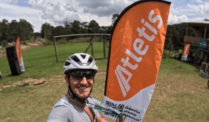 Selfie de um ciclista com bandeira da Atletis ao fundo, destacando um evento esportivo ao ar livre, ideal para promover atividades de atletas.