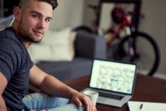 Homem sentado em um sofá olhando para a câmera, com um notebook aberto e uma bicicleta ao fundo.