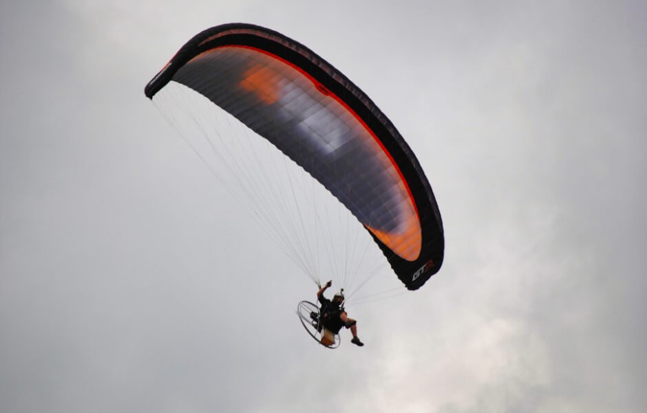 Imagem de um praticante de parapente em pleno voo, com a vela preta e laranja se destacando contra o céu.