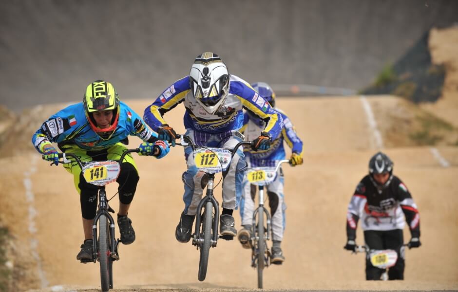 Imagem de um grupo de ciclistas participando de uma competição de BMX em uma pista de terra, usando capacetes e roupas esportivas coloridas.