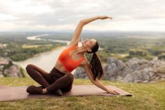 Mulher praticando yoga ao ar livre em uma paisagem natural com rio e montanhas ao fundo, realizando uma postura de alongamento sentado na grama.