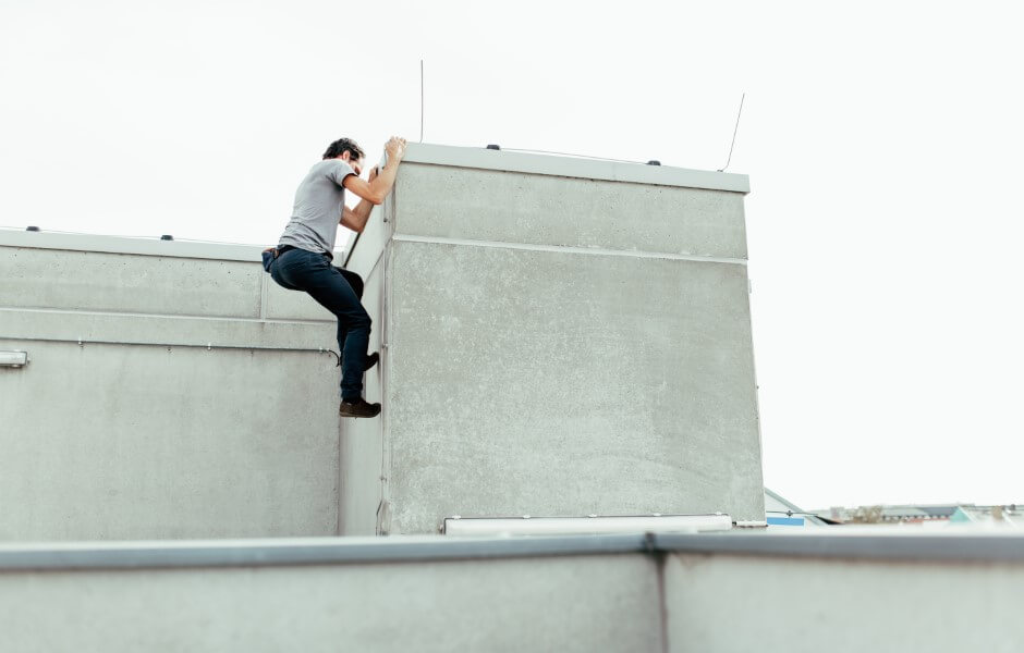 Imagem de um homem escalando um edifício, ele segura a beirada de uma parede com as duas mãos, enquanto apoia seus pés na parede.