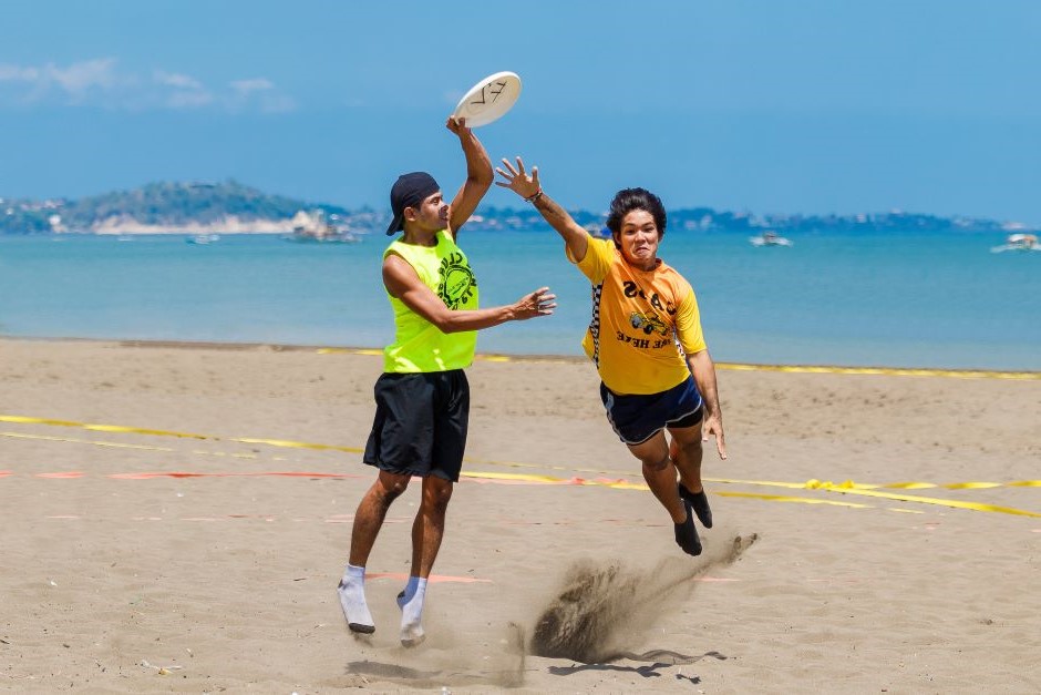 Dois atletas disputando o frisbee no ar dentro de quadra de areia à beira-mar