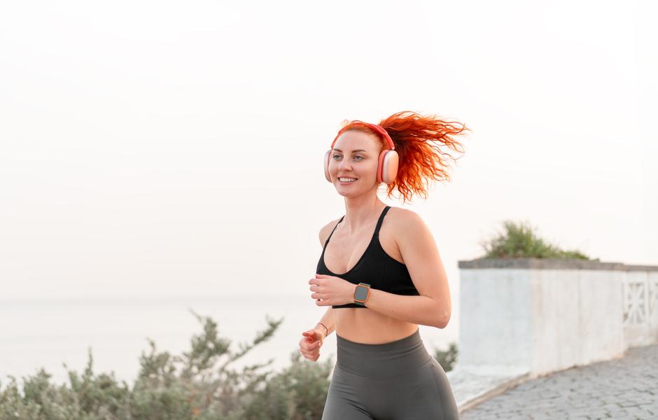 Mulher correndo ao ar livre com headphones, mostrando entusiasmo e energia. O cenário traz uma atmosfera tranquila para a prática de exercícios.