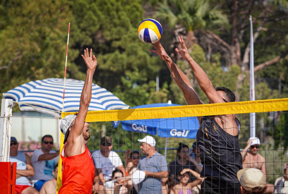 Dois jogadores homens de vôlei de praia disputando a bola na rede, no fundo da imagem arquibancada lotada de torcedores