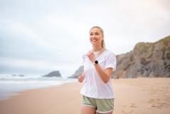 Mulher praticando corrida na praia, sorrindo e aproveitando o exercício ao ar livre com o mar ao fundo e uma paisagem de falésias ao longe.