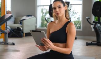 Jovem fitness em academia moderna, sentada no chão, sorrindo, usando roupa de ginástica preta, com tablet na mão, ambiente bem iluminado com equipamentos de treino ao fundo.