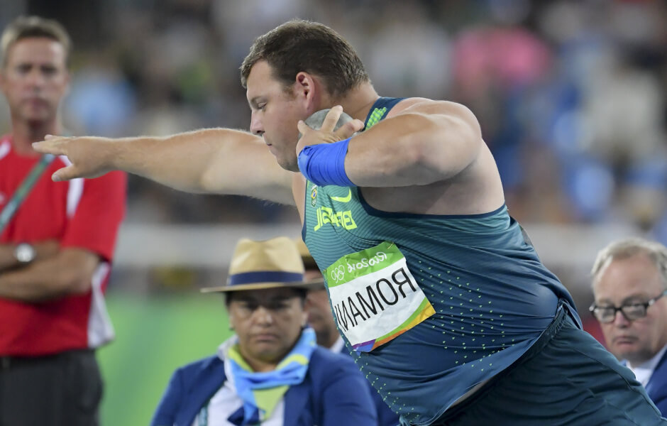 Imagem de um atleta com uniforme brasileiro segurando uma esfera de peso entre o ombro e o rosto, em posição de arremesso.