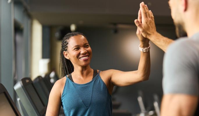 Mulher sorridente em treino na academia dando high five para seu treinador, demonstrando motivação e bem-estar na prática de exercícios físicos.
