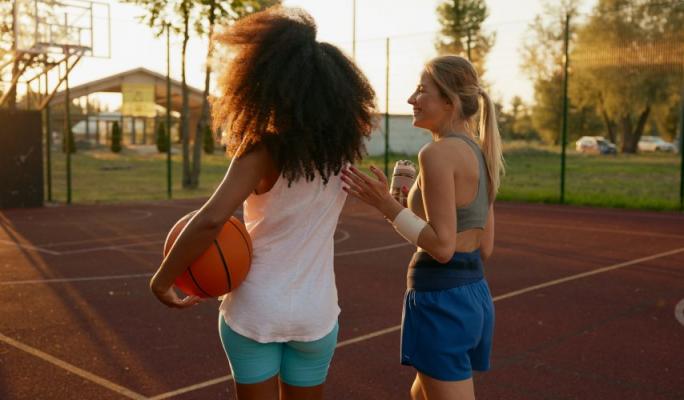 Duas jogadoras de basquete em uma quadra ao ar livre, uma segurando uma bola de basquete e a outra com uma bebida, desfrutando da prática esportiva durante o pôr do sol.