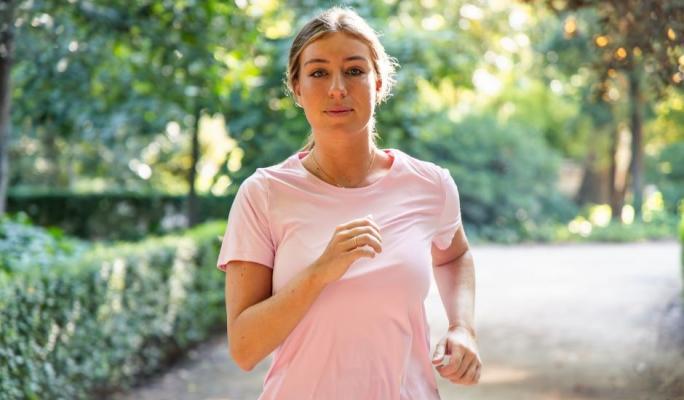 Mulher correndo ao ar livre em parque com árvores verdes na primavera, usando camiseta rosa, demonstrando saúde e bem-estar.