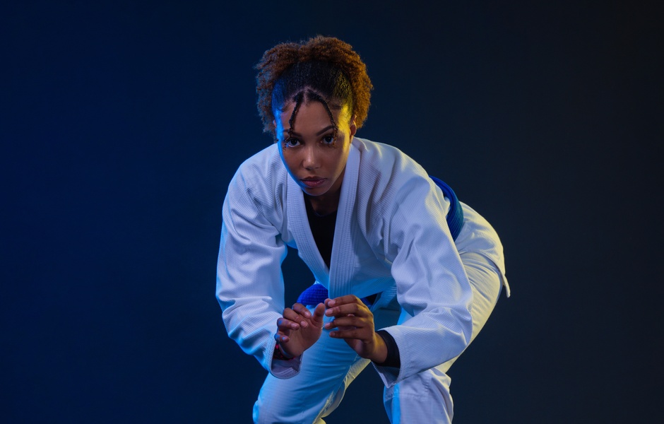 Atleta de jiu-jítsu em ação, utilizando um quimono branco, demonstrando técnica e foco em um ambiente de treino com fundo escuro e azul.