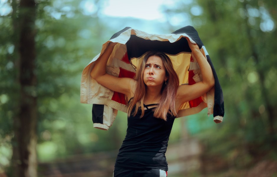 Imagem de uma mulher segurando uma jaqueta sobre a cabeça, presumivelmente para se proteger da chuva, enquanto está em uma floresta.