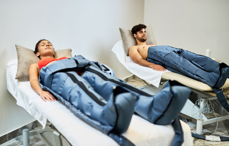 Foto de um homem e uma mulher deitados em camas separadas em uma sala de spa bem iluminada, ambos usando botas de compressão pneumática, relaxando com os olhos fechados.