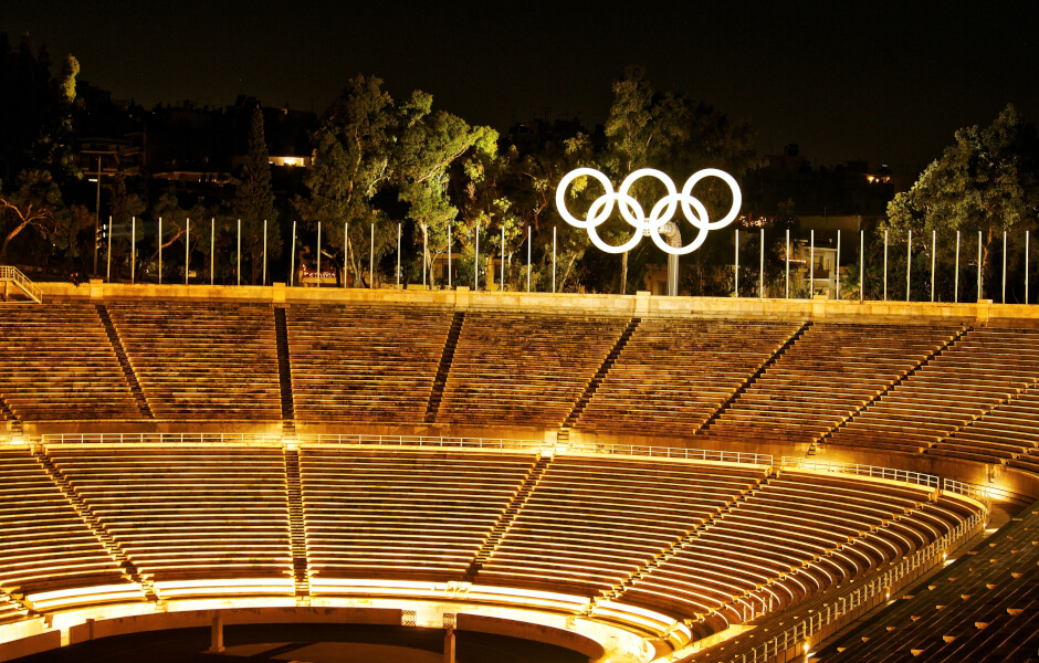 Imagem de uma grande arena com o logo das Olimpíadas. O ambiente é ao ar livre, à noite, e inclui um estádio com uma pista.