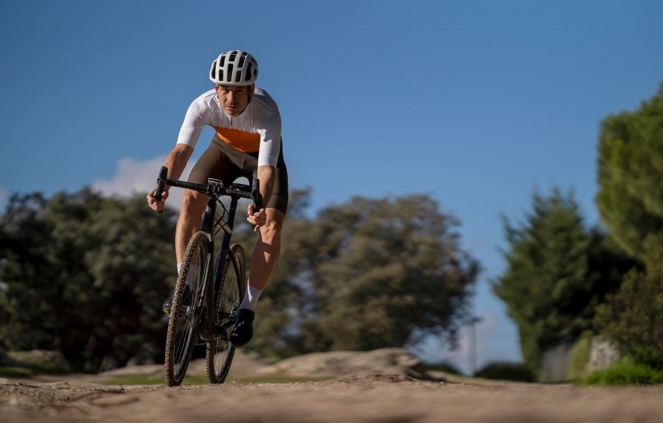 Homem praticando ciclismo off-road em ambiente natural ao ar livre com árvores e céu azul ao fundo, vestindo capacete e roupas esportivas.