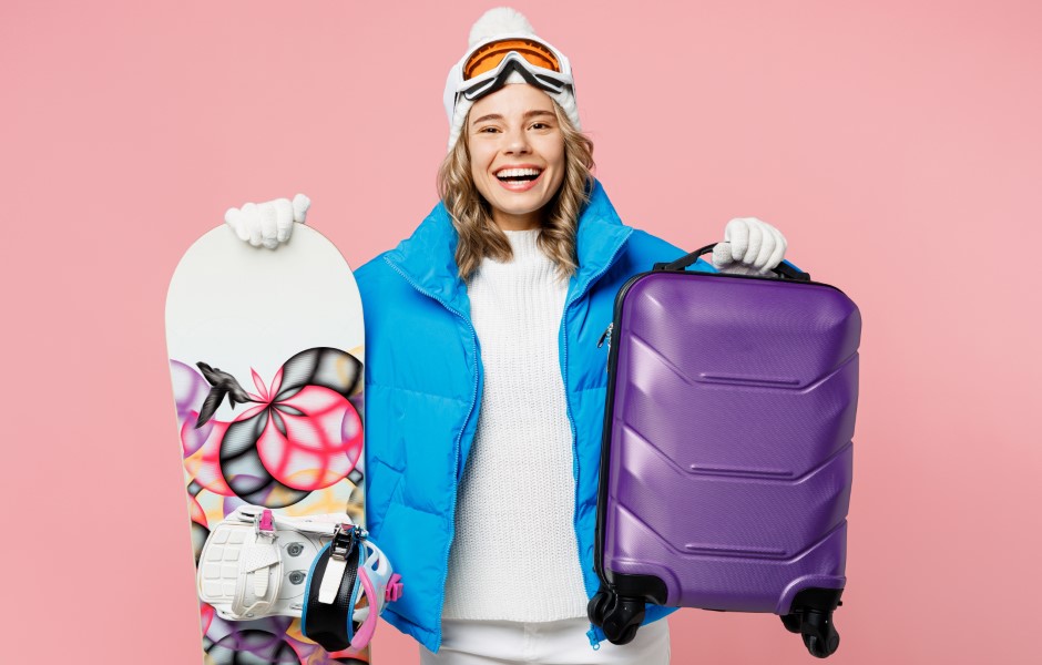 Imagem de uma mulher sorridente, vestida com roupas de inverno e óculos de esqui, segurando uma prancha de snowboard em uma mão e uma mala na outra.