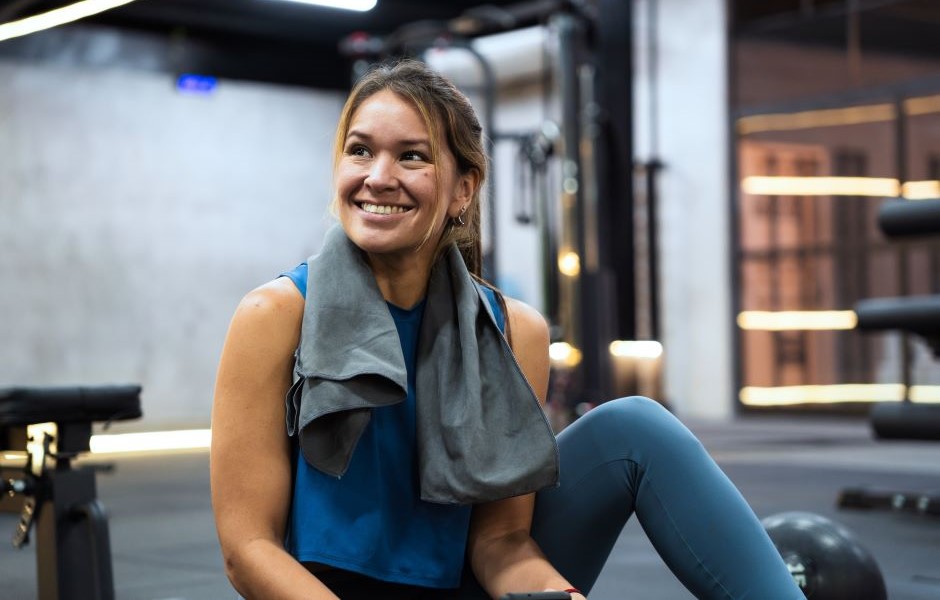 Mulher sorridente no supermercado com roupas esportivas, segurando um celular, após treino ou atividade física, ambiente moderno e bem iluminado.