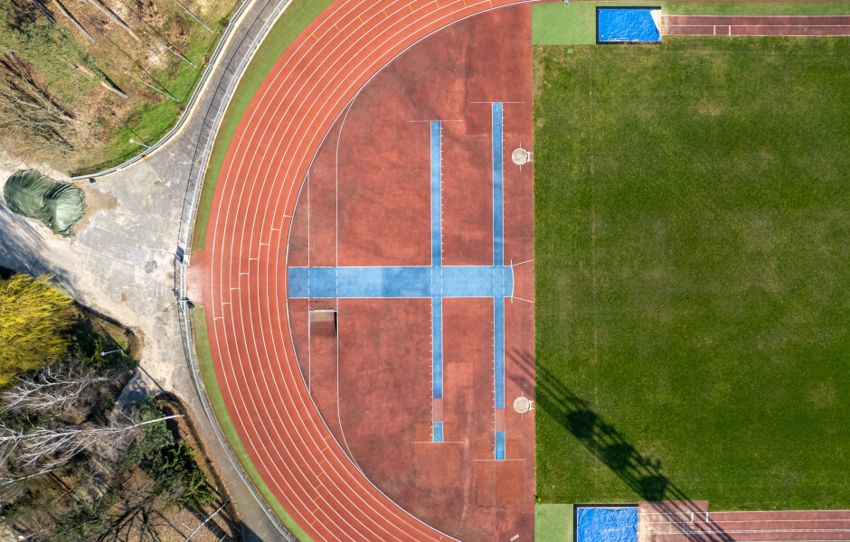 Imagem com uma visão aérea de uma pista de atletismo, que situa-se em volta de um gramado. A pista é laranja e possui linhas divisórias brancas, demarcando o trajeto dos atletas.