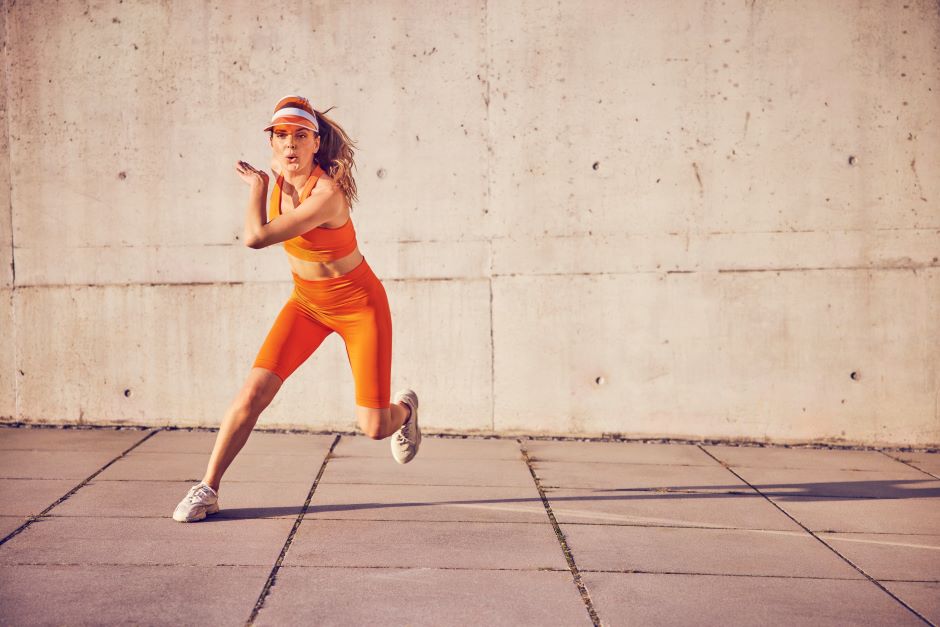Mulher se exercitando ao ar livre. Ela usa um conjunto de ginástica e uma viseira de cor laranja.