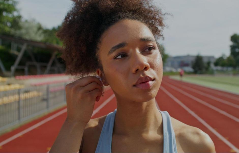 Mulher jovem negra com cabelo cacheado ajustando fone de ouvido sem fio enquanto se prepara para treinar em uma pista de atletismo vermelha ao ar livre.