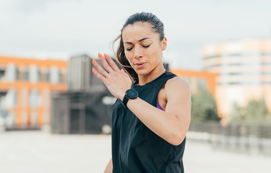 Mulher praticando corrida ao ar livre, verificando o relógio fitness, com expressão concentrada, em um ambiente urbano ao entardecer.