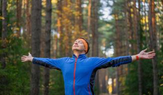Pessoa feliz se exercitando na floresta durante o outono, com braços abertos e expressão de bem-estar ao ar livre.