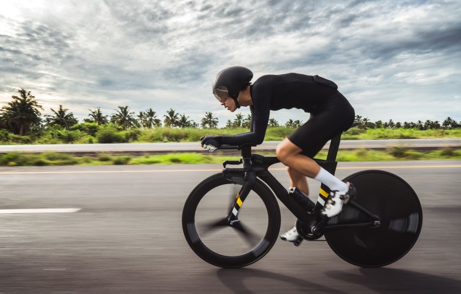Pessoa praticando ciclismo em alta velocidade na estrada com fundo de nuvens e vegetação verde ao redor, usando capacete e roupas esportivas.