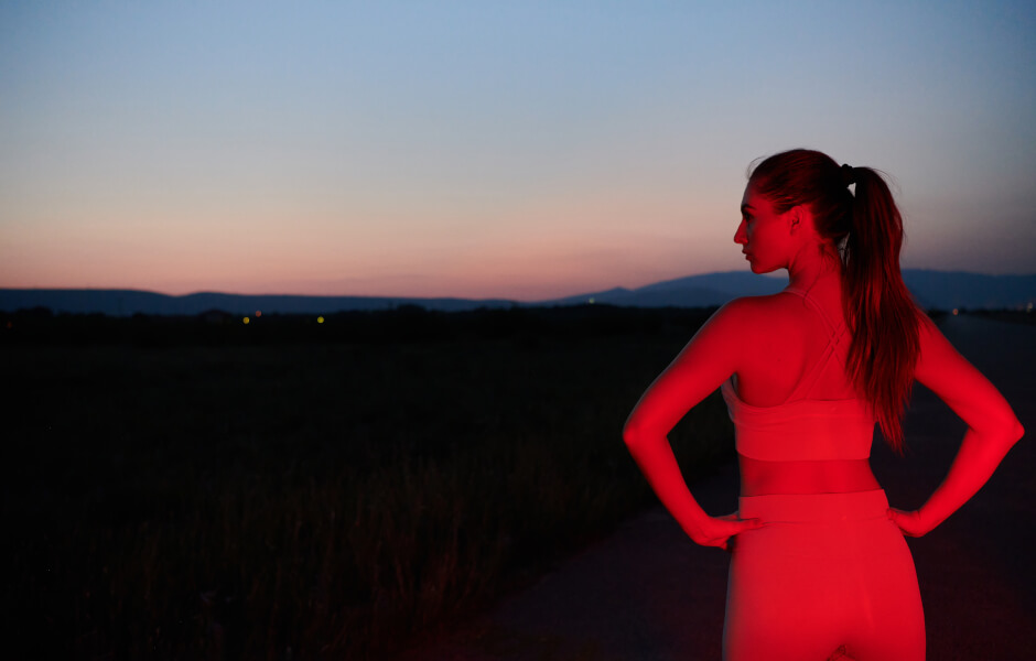 Imagem de uma mulher à noite, vestindo roupas de corrida. Ela está parada em pé observando bonita vista da cidade de cima de um morro.