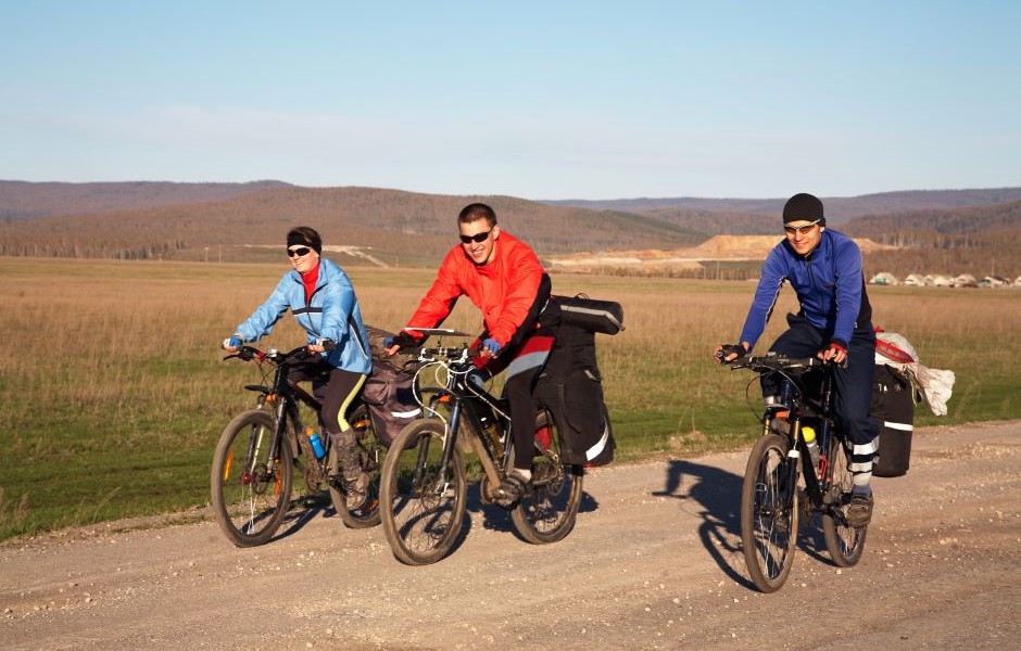 Três pessoas andam de bicicleta em um cenário de campo aberto, pedalando em uma estrada de terra sob um céu claro, aproveitando um passeio ao ar livre.