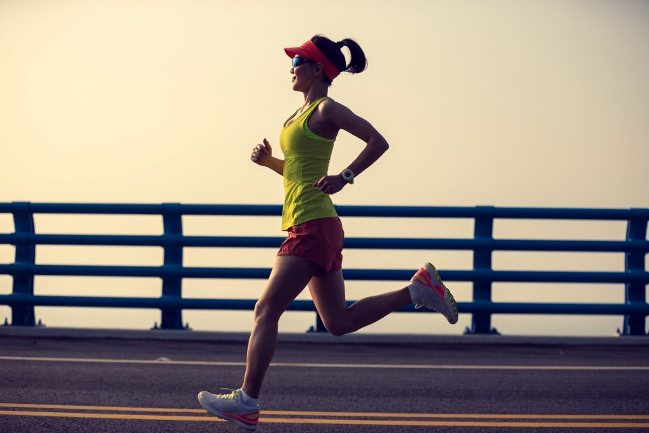 Imagem de uma mulher correndo em asfalto, com acessórios próprios para corrida. Ela usa, além de roupas específicas para corrida, óculos e viseira.