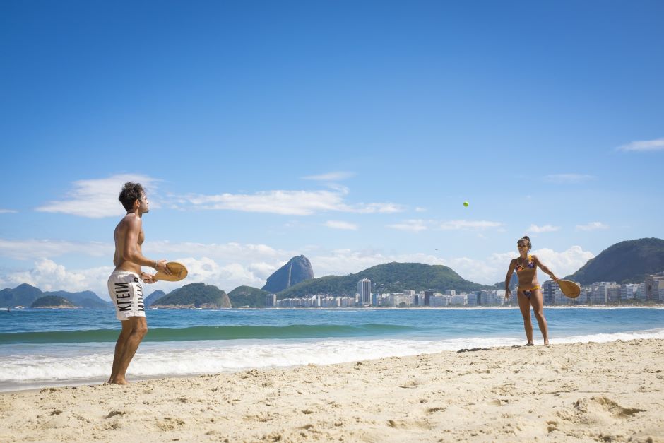 Dois jogadores, um homem com calção de banho e uma mulher com biquíni, praticando frescobol à beira-mar