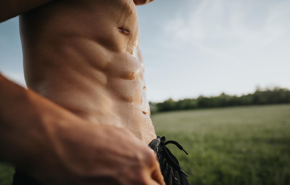 Imagem de um homem em forma exibindo seu abdômen definido em um ambiente externo.