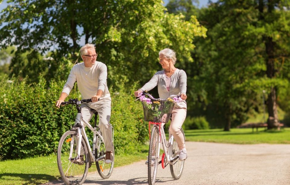 Casal de idosos pedalando em um parque sob árvores verdes, aproveitando a natureza, em um dia ensolarado, promovendo saúde e bem-estar na terceira idade.