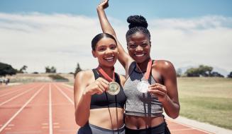 Duas atletas femininas sorridentes comemorando suas medalhas em uma pista de corrida ao ar livre, celebrando suas conquistas no esporte.
